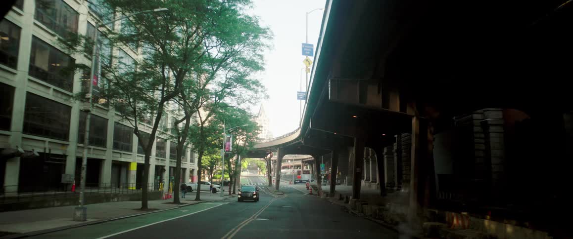 Vehicles drive down a city street beneath an elevated highway with buildings on one side and trees lining the sidewalk