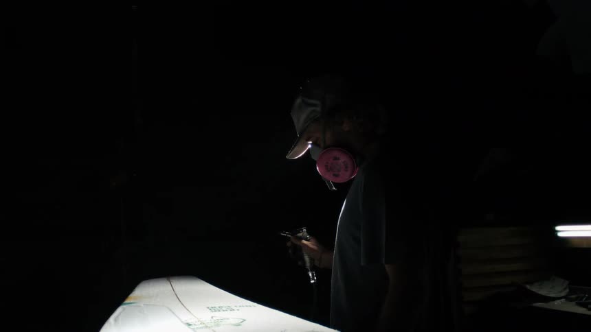 A person wearing a cap and respirator mask works on a surfboard in a dim room lit by a bright overhead light