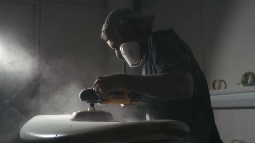 A carpenter wearing a face mask is using a smoothing tool to smooth out a surfboard inside the workshop