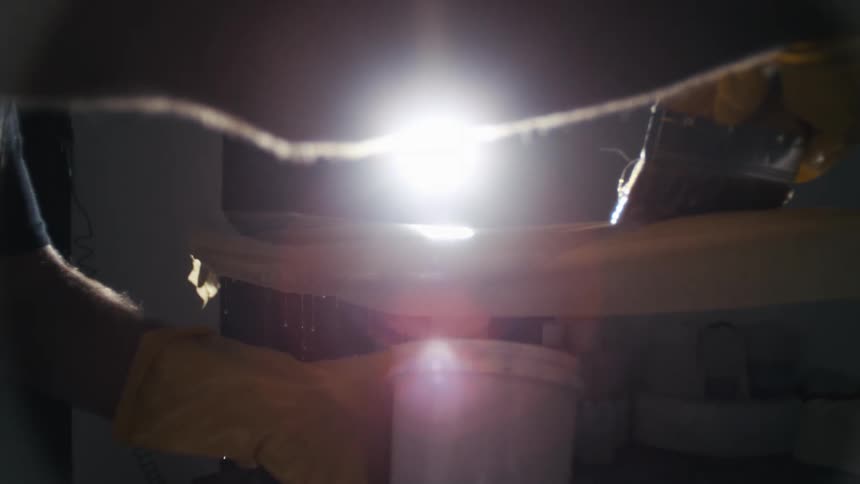The video shows hands wearing yellow gloves in a darkroom moving a piece of paper or film from a tray of liquid as the process continues a bright light flashes illuminating the scene and revealing liquid dripping into a white bucket