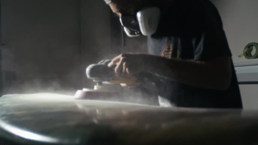 A carpenter wearing a face mask is using a smoothing tool to smooth out a surfboard inside the workshop
