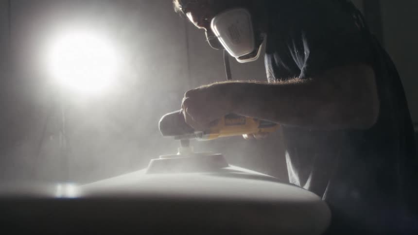 A person wearing a respirator and earmuffs uses a sander on a piece of material with dust flying in the air under a bright light