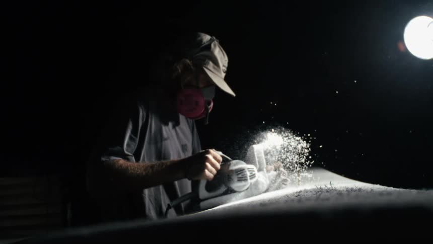 Man in cap and mask using an electric sander and polishing a surfboard