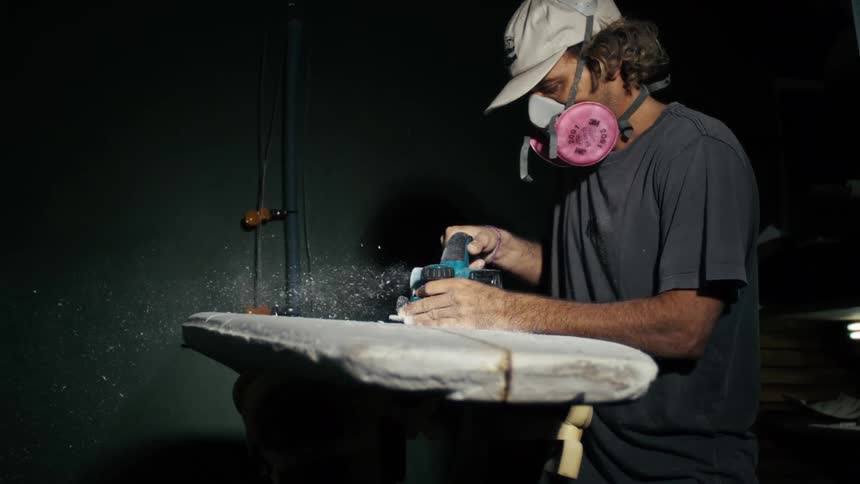 Concentrated man using an electric sandpaper and polishing a surfboard