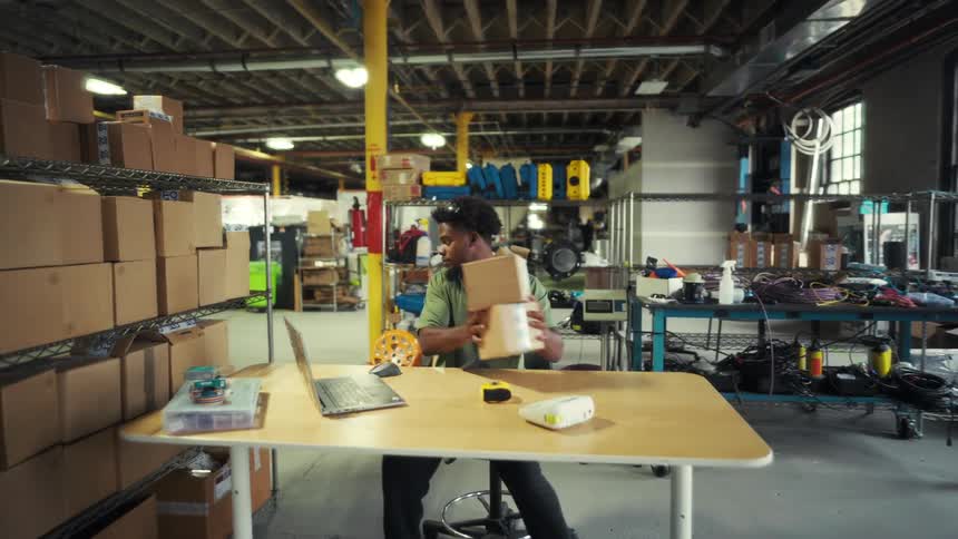 A young man is sitting at a desk inside a warehouse at midday preparing a shipping box and placing it on a shelf beside him with other shipping boxes a laptop is open in front of him