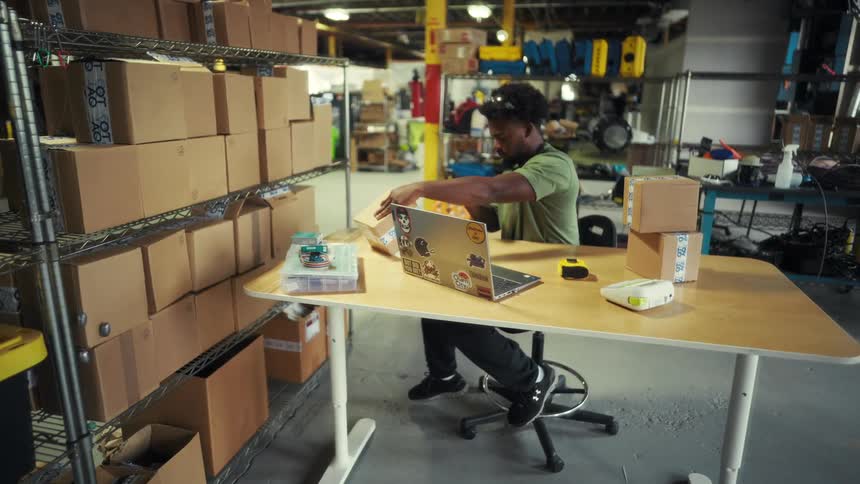 A person works at a laptop in a warehousestyle room filled with cardboard boxes organizing packages