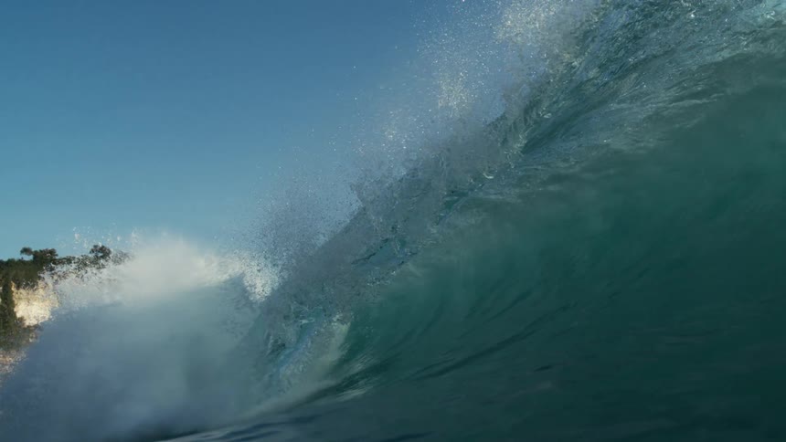 A large wave slowly folding over into the ocean under a blue sky