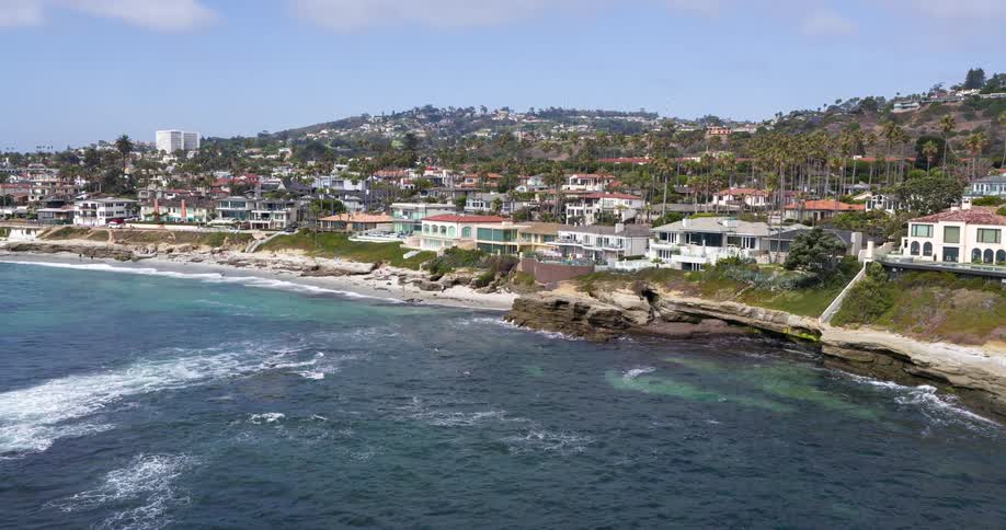 The video presents a scenic aerial view of a picturesque coastal community where luxurious houses with red roofs line a cliff overlooking the deep blue ocean lush green palm trees are scattered throughout the landscape enhancing the tropical feel