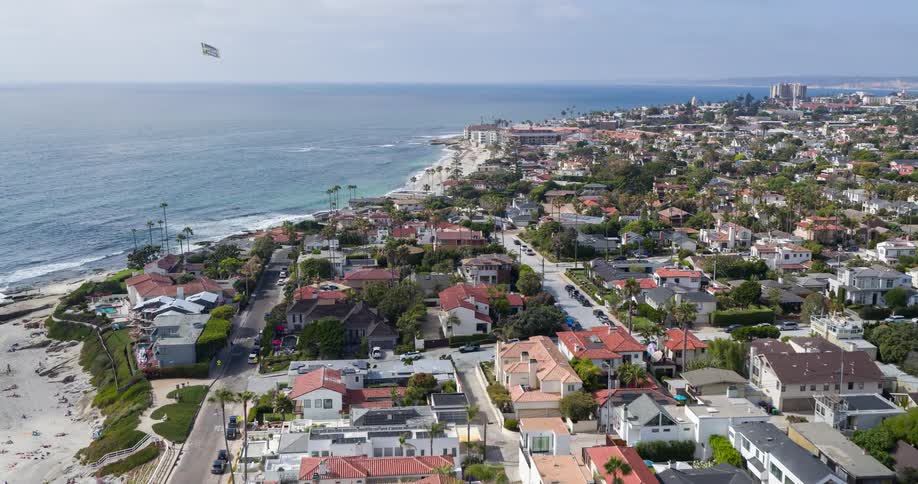 The video shows a scenic drone shot of a coastal town with numerous houses nestled on cliffs overlooking the ocean a long sandy beach stretches along the shoreline dotted with beachgoers enjoying the sunny weather