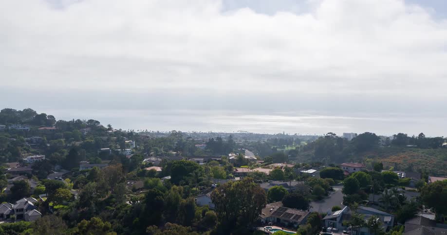 A quiet suburban neighborhood surrounded by dense trees stretches toward a cloudy ocean horizon