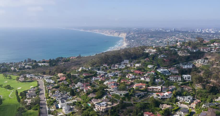 An aerial view of a residential neighborhood surrounded by vegetation with the city skyline and ocean visible in the background at midday