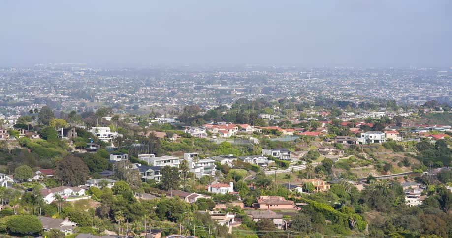 A broad view of suburban neighborhoods extending across a green hilly landscape under a hazy sky