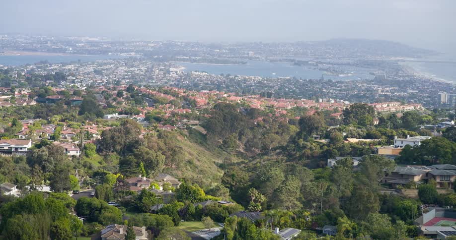 The video provides a highangle sweeping view of a vast urban landscape dotted with numerous residential properties and lush green trees in the distance a large body of water likely a bay or ocean is visible under a somewhat hazy sky