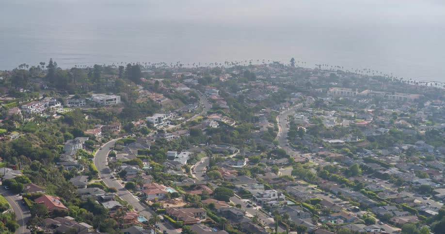 The video provides an expansive aerial view of a sprawling coastal city with numerous houses nestled on hillsides a hazy sky hangs over the horizon where the land meets a large body of water likely a bay or ocean