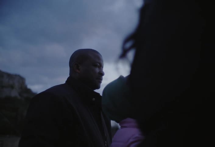 A family stands outdoors at twilight with an adult in a jacket beside a girl wearing a green beanie decorated with colorful beads