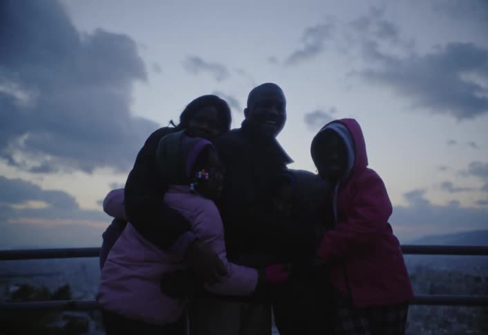 A family of five is standing on the side of the road beside a fence rail sharing a group hug as a car passes by at sundown with the cityscape visible in the background