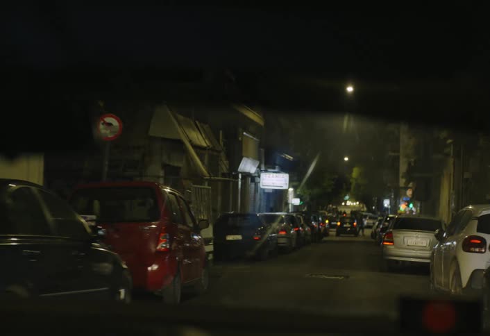 A dimly lit urban street lined with parked cars seen through a vehicle windshield at night