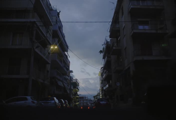 A vehicle drives down a street lined with apartment buildings at dusk with other cars parked and driving on the road