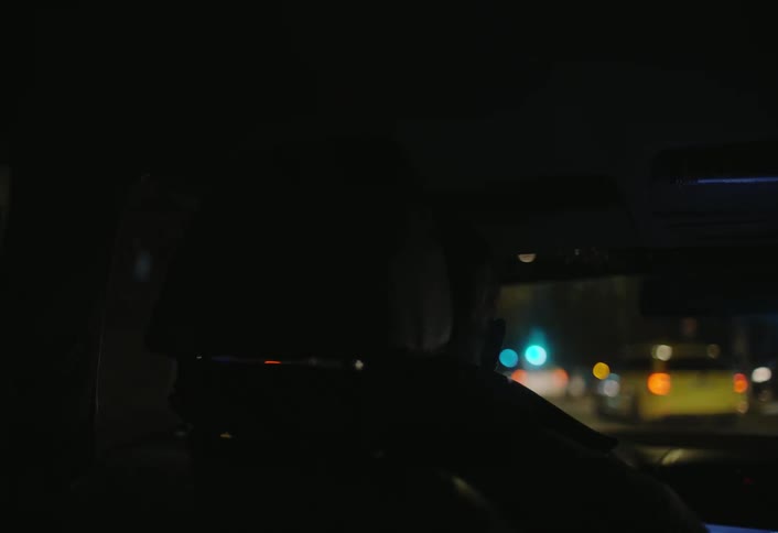 A person is driving a car at night viewed from the back seat with the lights of other vehicles and the city blurred in the background