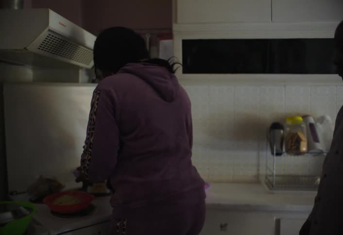 A woman prepares food in a modest kitchen with bowls and utensils scattered across the counter