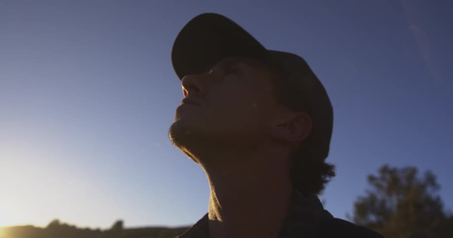 A young man wearing a hat looks up at the sky as the sun sets in the woods