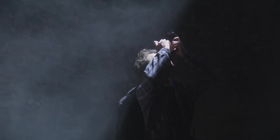 A man stands in a beam of light looking up through a viewfinder in a dark smoky room