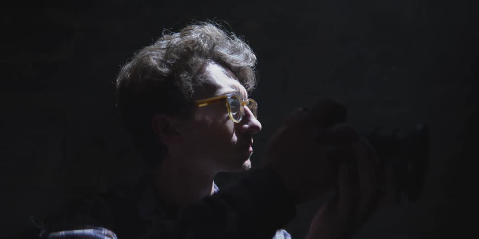 A man looks through the viewfinder of a camera framed dramatically in low light