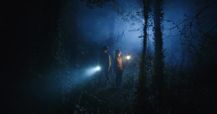 Pair of teenagers in misty forest at night looking around with a flashlight and spotlight