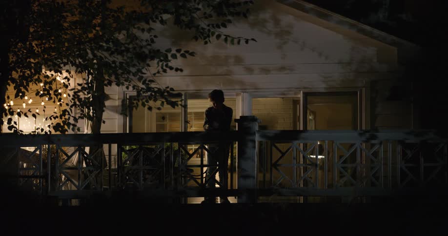 A teenager stands silhouetted on a porch at night checking his smartphone as warm indoor light spills through the doorway