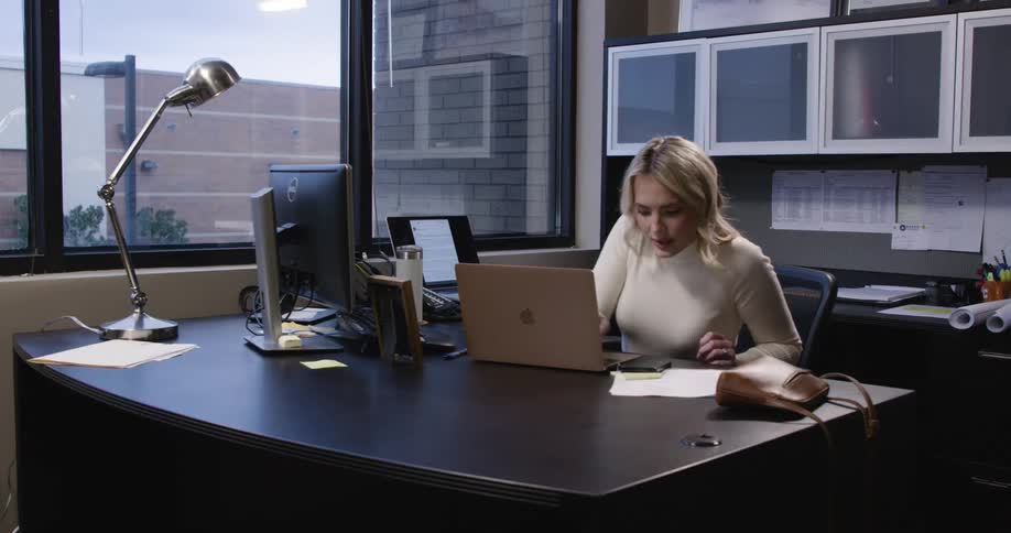 A woman works at a desk in a small office surrounded by a desktop monitor laptop lamp and scattered supplies