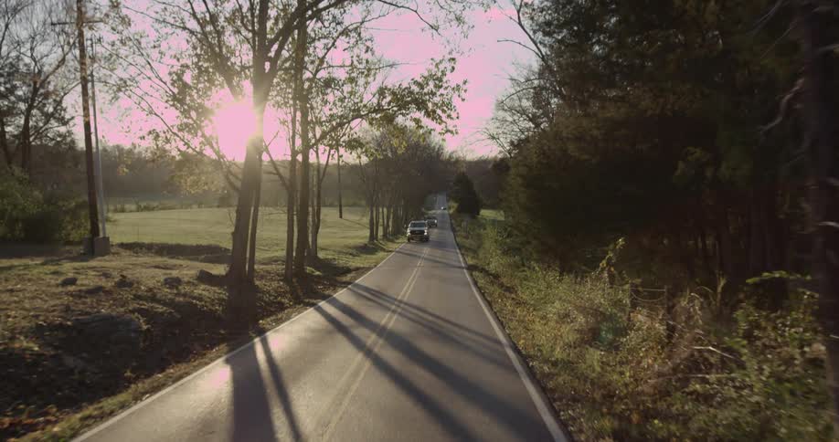 Vehicle traffic on a narrow street surrounded by fields and trees