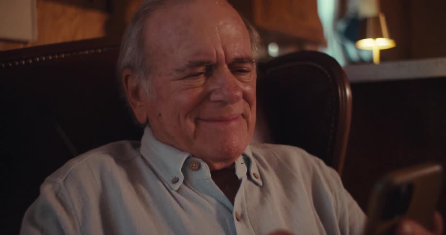 The video shows an elderly man sitting in a comfortable leather chair engrossed in his smartphone he appears to be smiling or amused by something on the screen