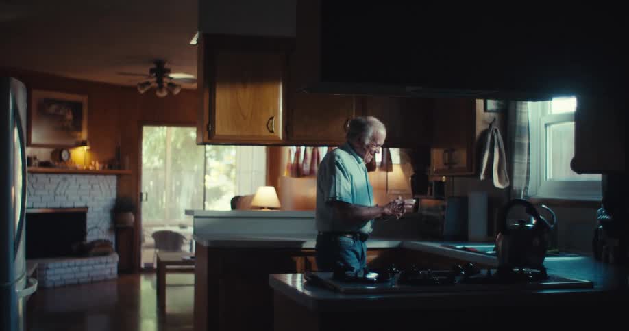 Older man stands in a dimly lit kitchen smiling while looking at his phone