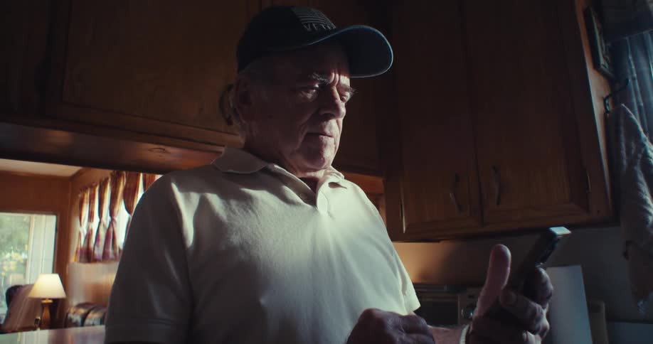 Elderly man wearing a cap in the kitchen holding and interacting with a smartphone