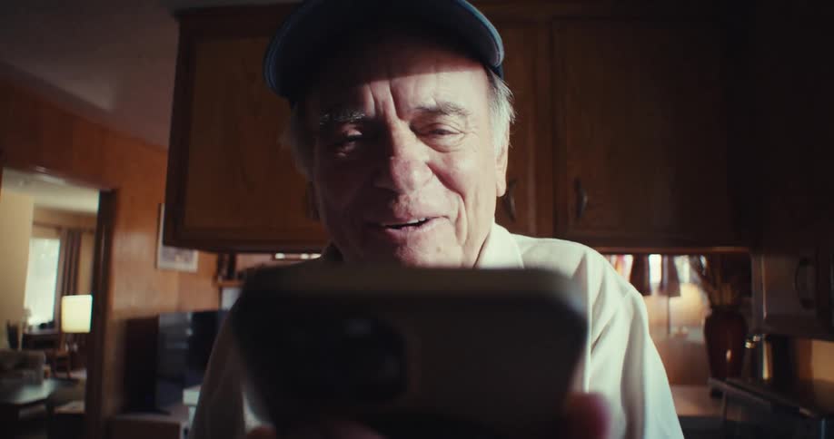 The video features a closeup shot of an elderly man holding a smartphone he is wearing a blue baseball cap and appears to be engaged in a video call or recording a message as he smiles and waves at the camera