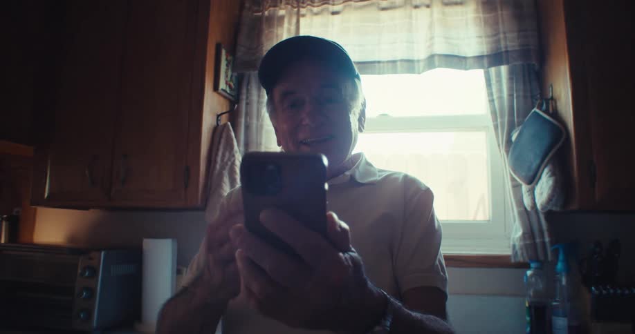 Elderly man standing in his kitchen in front of window facetiming on his cell phone