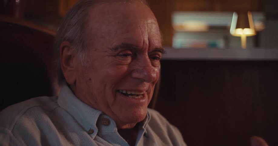 The video features an elderly man sitting in a comfortable chair wearing a lightcolored collared shirt he appears to be talking and gesturing possibly on a video call or telling a story