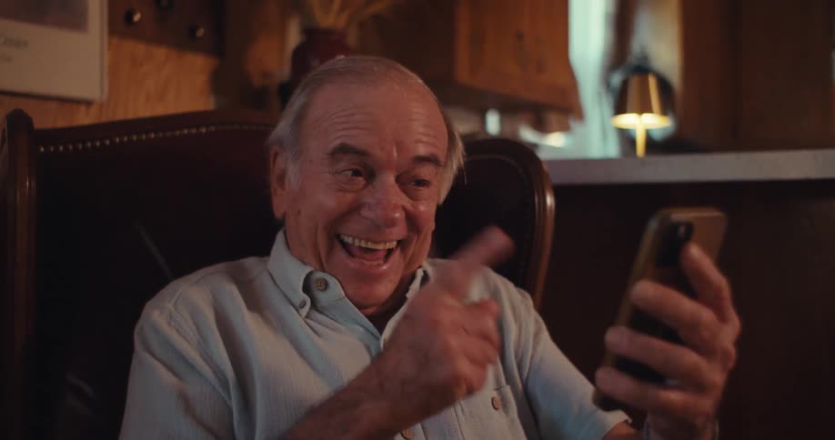 An older man with light colored hair and a light blue collared shirt smiles broadly while looking at a cell phone in his hands