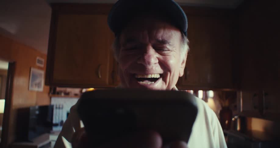 An elderly man happily video chats on his phone in a cozy kitchen