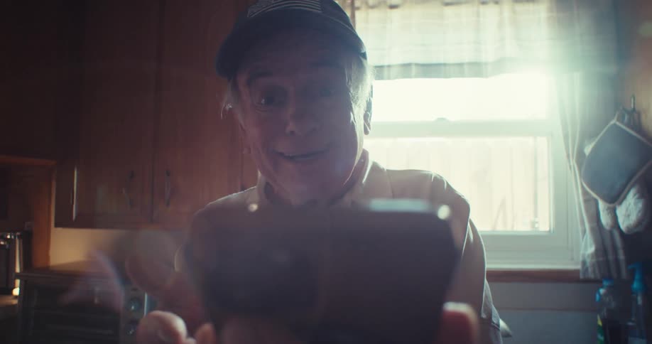 Glaring sun shines through the window behind an elderly man while he is surprised and pleased by the contents displayed on his smartphone