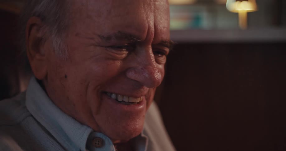 The video features a blurred closeup of an elderly man holding a smartphone with a veteran baseball cap visible he appears to be talking and laughing while looking at the screen with sunlight creating a glare in the background