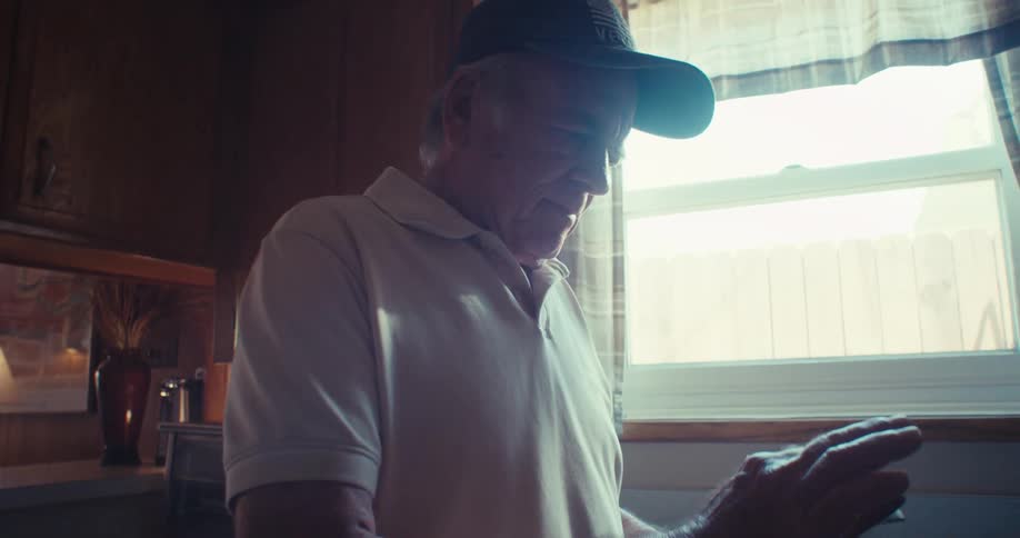 An elderly man is standing in the kitchen beside the sink at midday wearing a hat and scrolling through his smartphone