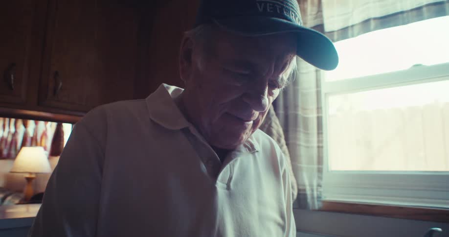 The video shows a man wearing a baseball cap in what appears to be a kitchen or dining area illuminated by bright light from a window he is focused on his phone which he holds in his hands