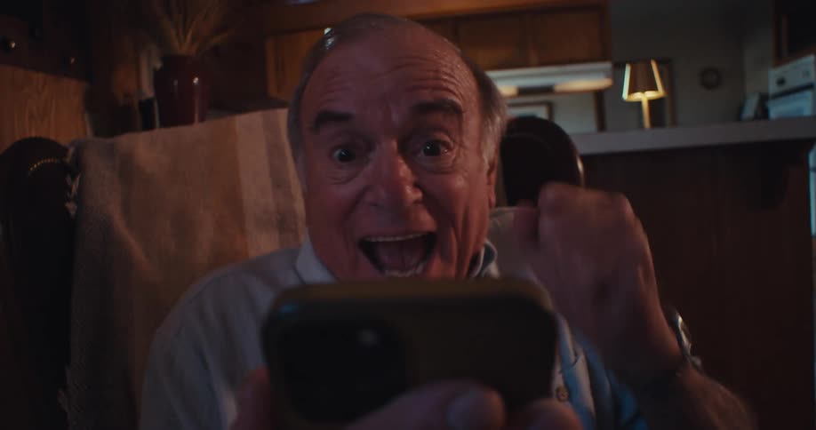 The video features a closeup shot of an elderly man holding a smartphone looking at the camera with an expression of surprise that quickly turns into a wide smile and laughter he appears to be reacting to something he sees on the phones screen