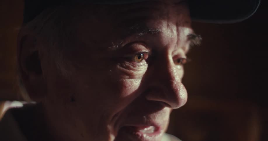 An elderly man standing and talking indoors wearing a baseball hat