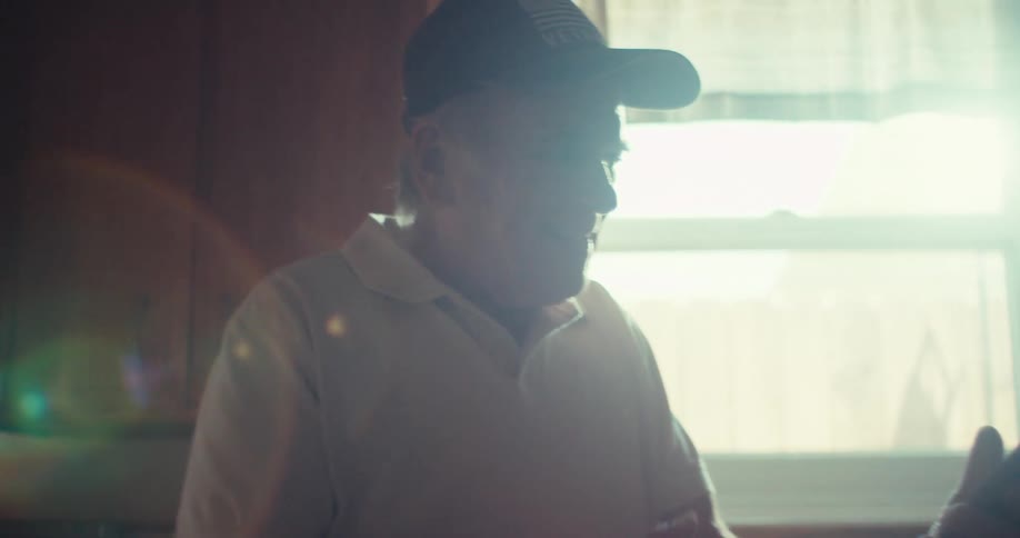 The video shows a man from the side wearing a cap and a lightcolored shirt with sunlight streaming from a window in the background he appears to be talking or interacting with someone or something offcamera