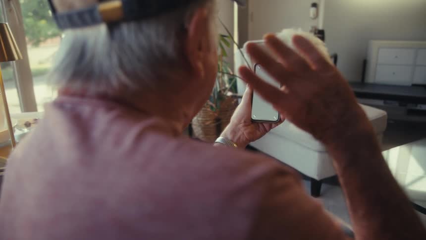 Elderly man sitting in his living room waves while making a video call on his smartphone