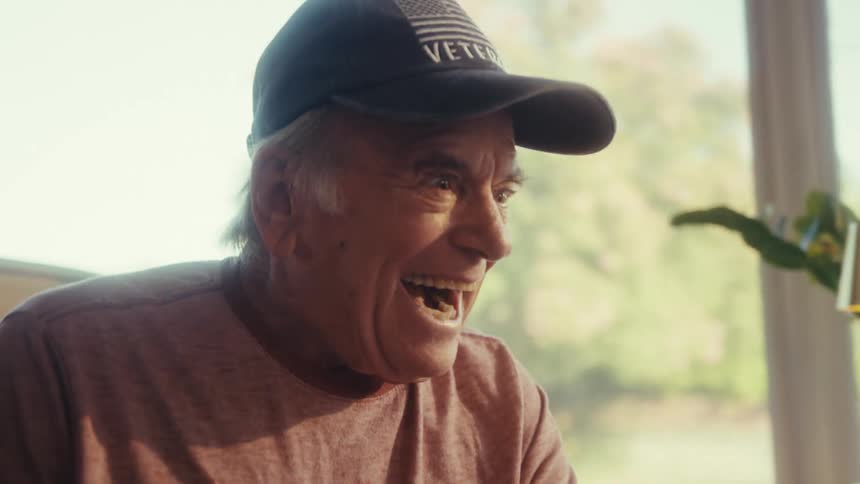 A man wearing a veteran cap and a pink shirt reacts with exaggerated surprise and then laughter while looking at a smartphone