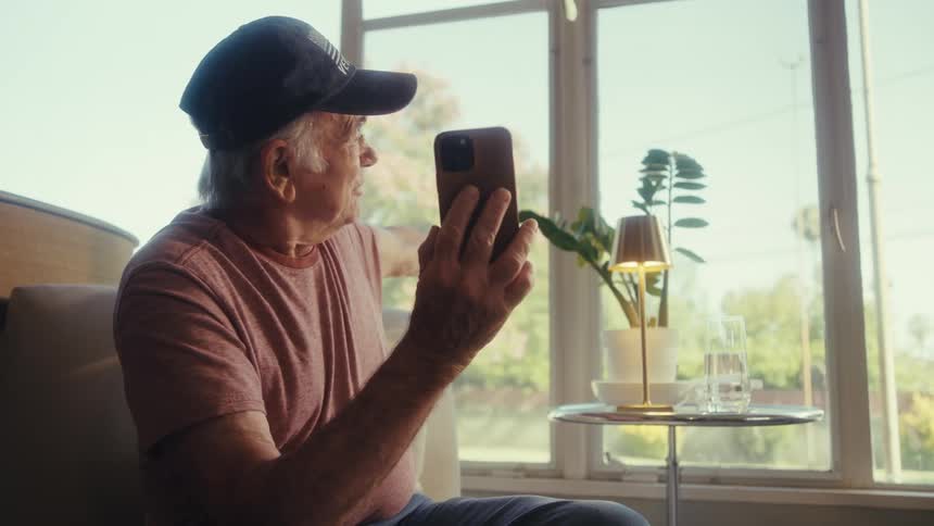 An elderly man sitting on a chair smiling and talking to someone on a video call holding his phone in one hand while pointing out the window