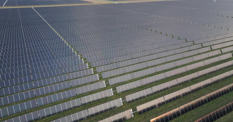 Twisting traverse over an expansive solar farm with numerous rows of gleaming solar panels extending across a green field